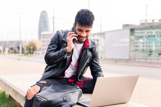 smiling latin man typing on his computer while talking on the phone sitting in the city, concept of technology and urban lifestyle