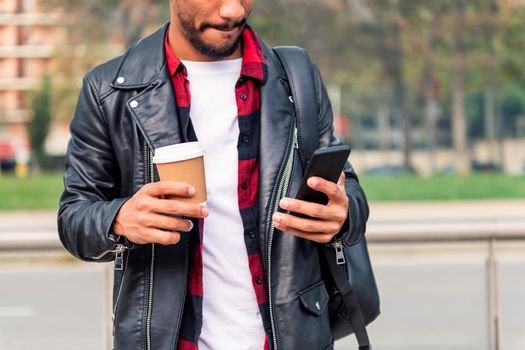 detail of the hands of a stylish young latin student consulting his phone with a take away coffee, concept of technology and urban lifestyle