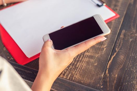 Mockup image of a woman's hands holding white mobile phone with blank black screen on background of white sheet of paper at a cafe.