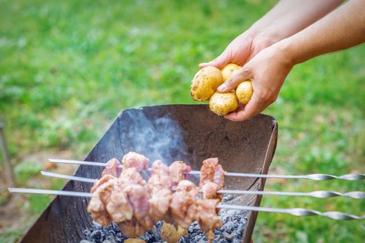 Hands of man prepares barbecue meat with potatoes on skewer by grill on fire outdoors. Concept of lifestyle rustic food preparation