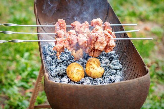 Hands of man prepares barbecue meat with potatoes on skewer by grill on fire outdoors. Concept of lifestyle rustic food preparation