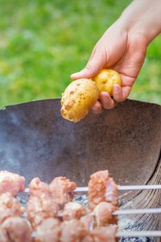 Hands of man prepares barbecue meat with potatoes on skewer by grill on fire outdoors. Concept of lifestyle rustic food preparation