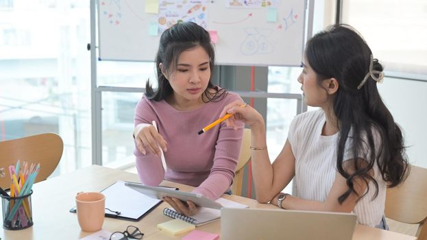 Two young asian women sitting at office desk and talking about project startup ideas.