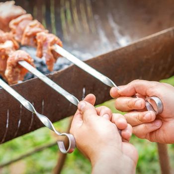 Hands of man prepares barbecue meat on skewer by grill on fire outdoors