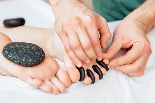 Male hands putting hot little black pebbles between female toes for feet massage in spa salon