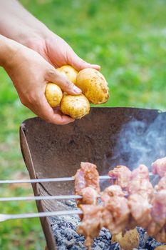 Hands of man prepares barbecue meat with potatoes on skewer by grill on fire outdoors. Concept of lifestyle rustic food preparation