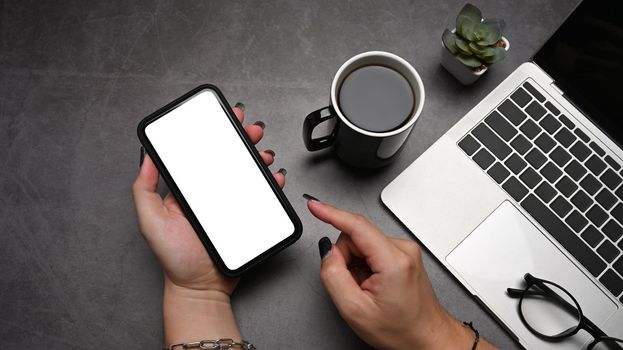 Woman hand holding mobile phone with blank screen on dark stone background. Top view.