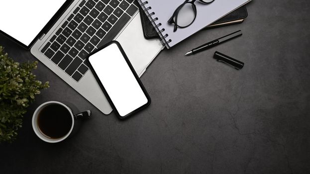 Mobile phone with blank screen, laptop computer, coffee cup and eyeglasses on dark stone background. Top view.
