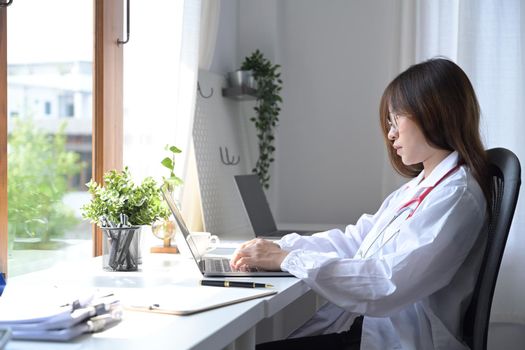 Side view female doctor in white uniform using laptop computer at medical office.