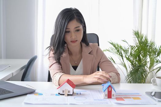 Millennial businesswoman sitting in office with house model. Accounting, property investment concept.