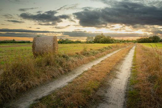 Country road and hay bale in the field, evening view with clouds, Nowiny, Poland