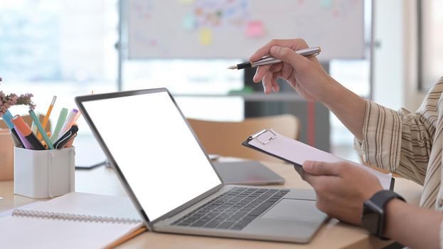 Male office worker holding clipboard and using laptop computer.