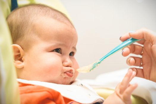 Baby in high chair does not want to eat food that mother gives with spoon, naughty and crying. Concept of baby food, introduction of complementary foods. Selective focus. High quality photo