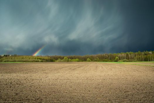 Fantastic clouds with a rainbow and a ploughed field, spring rural view