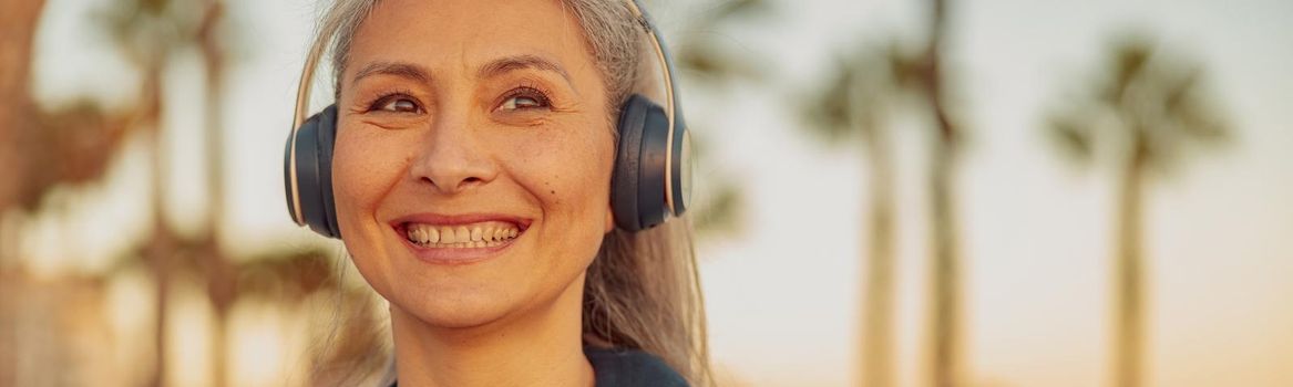Smiling mature woman listening to music in hedphones standing on background of seafront with palm trees, looking to the side