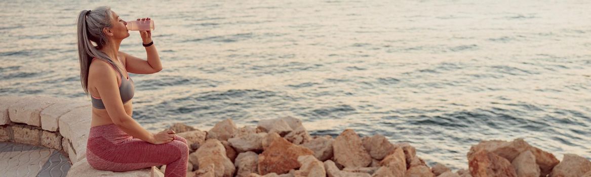 Asian lady in sportswear sitting on the edge of the coastal fence, drinking water, on background with blue sea and sunset