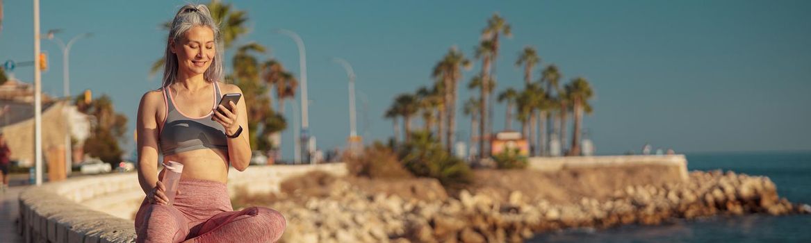 Active woman in sports clothes sitting by the seashore, holding mobile phone and lookind at it