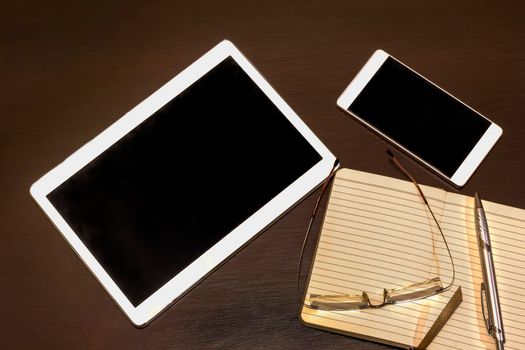 Office table: mobile phone, tablet, glasses, notebook and pen with copy space. Gadgets on a wooden table. Working place, top view