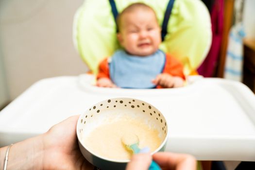 Baby in high chair does not want to eat food that mother gives with spoon, naughty and crying. Concept of baby food, introduction of complementary foods. Selective focus. High quality photo