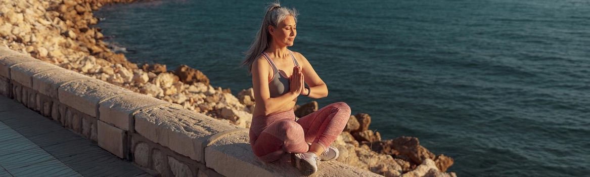 Pretty adult female in sports top and leggings resting by the seashore in lotus position and taining on sunny day