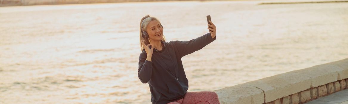 Beautiful Asian lady holding smartphone and taking picture of herself while sitting on waterfront on the backgroung of calm sea