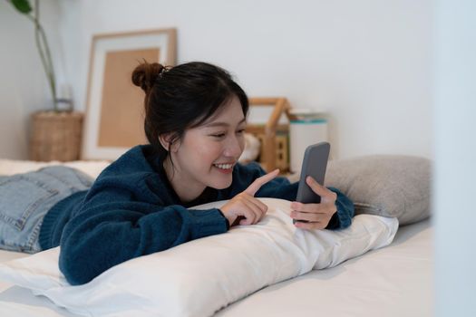Beautiful asian woman laying down on white bed using mobile phone and smile in morning sunday