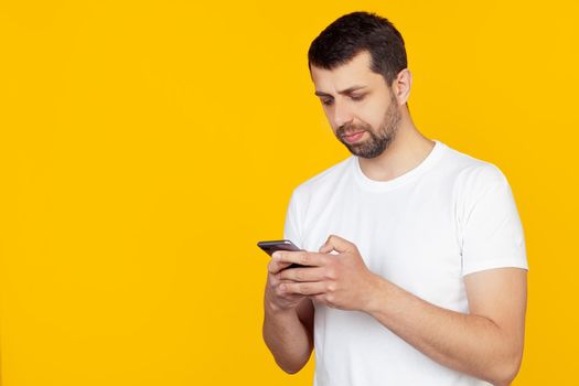 A young man with a beard in a white t-shirt uses a smartphone with a confident expression on his smart face, thinking seriously. Stands on isolated yellow background.