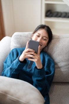 Image of smiling attractive asian woman using mobile phone while lying on sofa at home.