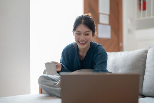 Happy young asian woman using laptop computer on cozy sofa, sunday morning concept.