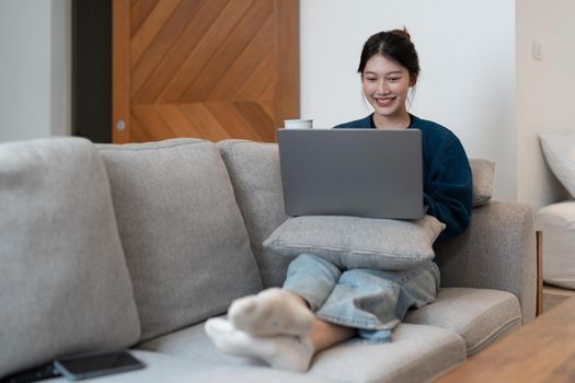 Happy young woman in a blue sweatshirt works at home on a blue sofa with a laptop and a cat, remote work and education, staying home during a virus.