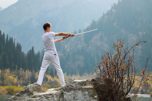 A young man stands on a large stone and holds a Japanese sword in his hands