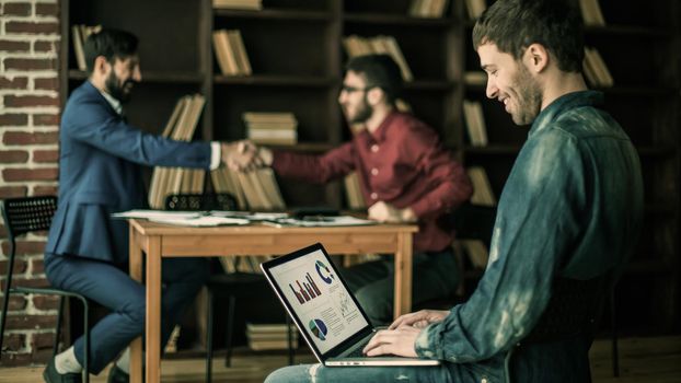 sales Manager working on laptop with a marketing report on the background of business partners handshaking