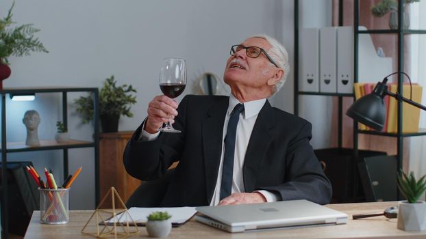 Senior man freelancer drinking wine from glass after working on laptop computer pc sitting at desk at home office. Elderly mature guy e-learning, online distant video call conversation remote job