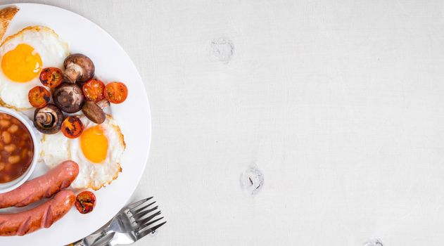 Full english breakfast with fried eggs, tomatoes, sausages, bacon, mushrooms, toasts and beans. Breakfast on a white plate with forks on the white wooden table. Space for text. Background