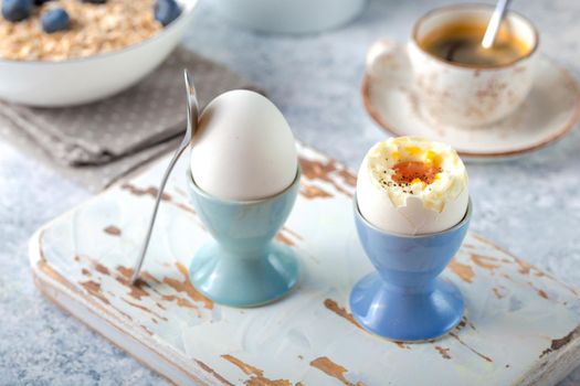 Fresh soft boiled eggs, oatmeal with blueberries, coffee cup, milk jug. White concrete rustic background. Soft eggs, healthy breakfast. Selective focus. Tasty light fitness breakfast concept. Closeup