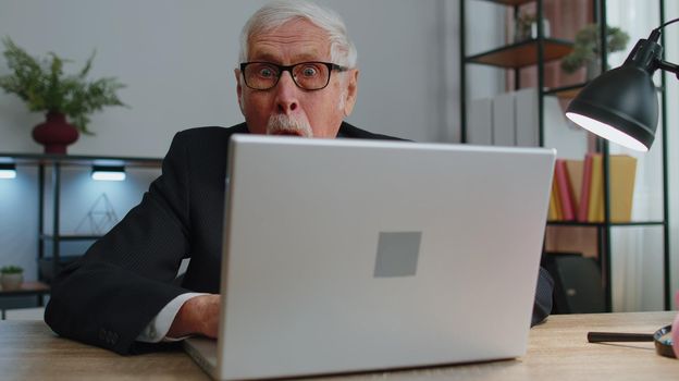 Senior mature older business man hiding behind laptop computer, making funny face, fooling around, putting her tongue out remote working disrespecting someone at home office. One elderly grandfather