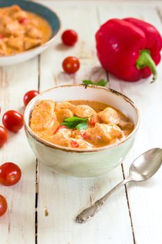 Delicious chicken stew with paprika in a bowl on a white wooden table. With fresh cherry tomatoes, red bell pepper and parsley. Traditional hungarian dish paprikash. Comfort food. Selective focus