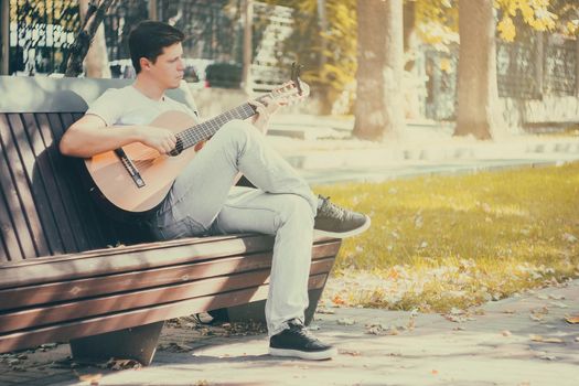 Young man sitting on the bench in the park playing on acoustic guitar with capo. Young attractive man enjoys live music in last sunny days autumn holiday. Retro lens used.