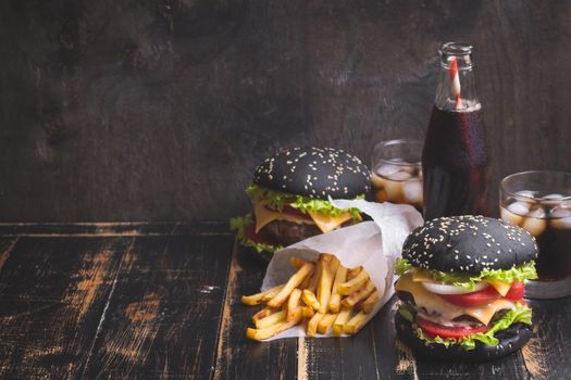 Set of black burgers with meat patty, cheese, tomatoes, mayonnaise, french fries and glass of cold cola soda with ice. Dark wooden rustic background. Space for text. Modern fast food. Toned image