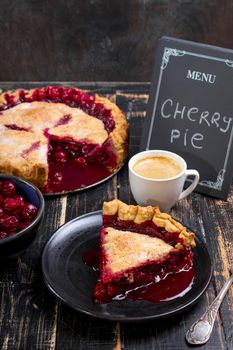 Slice of homemade cherry pie, cup of coffee, bowl with cherries and menu chalkboard on the black wooden table