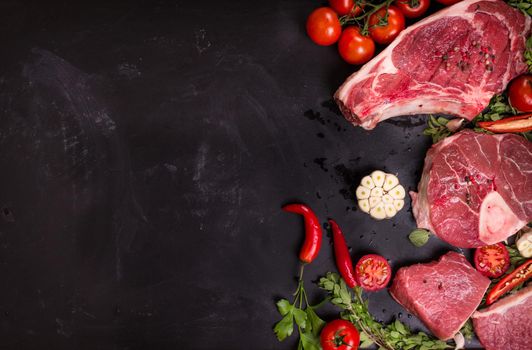 Raw juicy meat steaks ready for roasting on a black chalk board background. Rib eye steak on the bone, veal shank (ossobuco), fillet with cherry tomatoes, hot pepper and herbs. Space for text