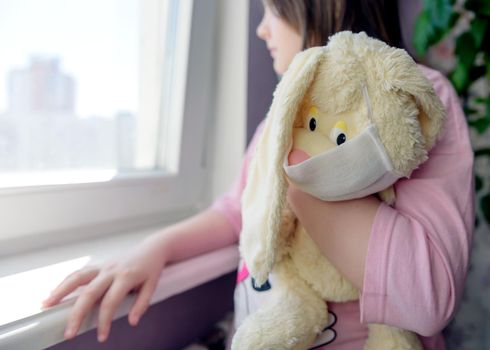 A girl with her toy, a plush rabbit, in a mask looks out the window concept of protection against coronavirus, quarantine, stay at home.