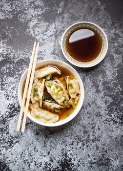 Asian dumplings in broth, bowl, chopsticks, soy sauce, rustic stone background. Top view. Chinese dumplings for dinner. Closeup. Traditional Asian/Chinese cuisine. Overhead. Soup