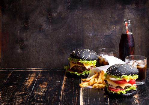 Set of black burgers with meat patty, cheese, tomatoes, mayonnaise, french fries and glass of cold cola soda with ice. Dark wooden rustic background. Space for text. Modern fast food lunch frame