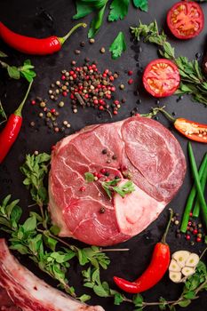 Raw veal shank (ossobuco), on a black chalk board background with cherry tomatoes, hot pepper and herbs. Ready for cooking