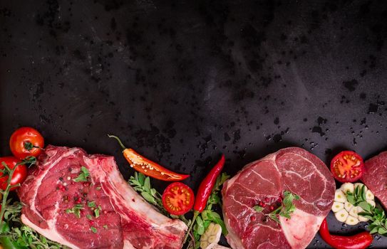 Raw juicy meat steaks ready for roasting on a black chalk board background. Rib eye steak on the bone, veal shank (ossobuco), fillet with cherry tomatoes, hot pepper and herbs. Space for text