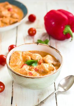 Delicious chicken stew with paprika in a bowl on a white wooden table. With fresh cherry tomatoes, red bell pepper and parsley. Traditional hungarian dish paprikash. Comfort food. Selective focus