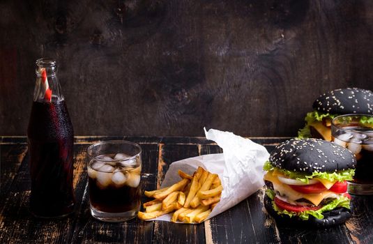 Set of black burgers with meat patty, cheese, tomatoes, mayonnaise, french fries and glass of cold cola soda with ice. Dark wooden rustic background. Space for text. Modern fast food lunch frame