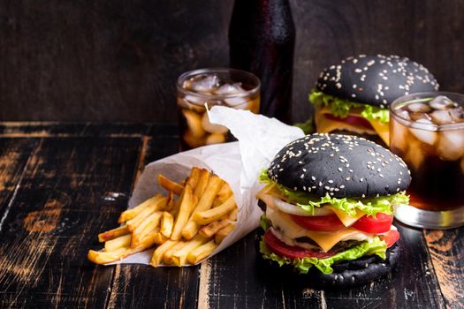 Set of black burgers with meat patty, cheese, tomatoes, mayonnaise, french fries in a paper cup and glass of cold cola soda with ice. Dark wooden rustic table. Modern fast food lunch