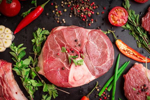 Raw veal shank (ossobuco), on a black chalk board background with cherry tomatoes, hot pepper and herbs. Ready for cooking